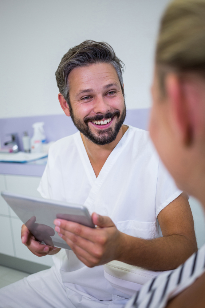 A smiling male dentist in white scrubs holds a tablet while talking to a patient.